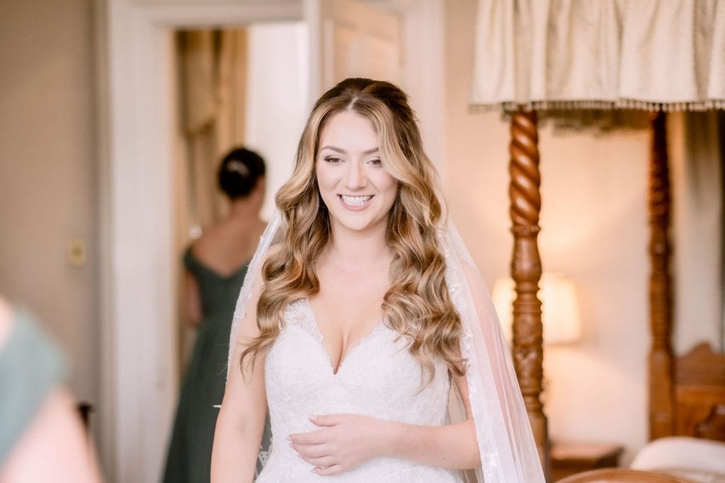 A smiling bride in a white dress and veil, standing in a warmly lit room at Orchardleigh House, with a bridesmaid visible in the background.