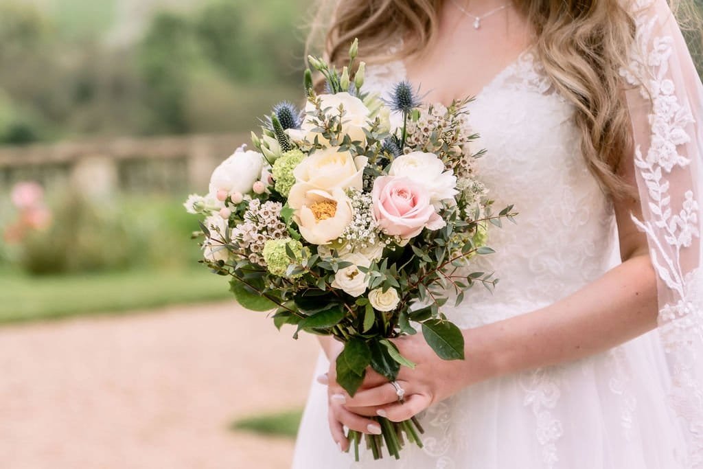 Bride in a lace dress holding a bouquet of white and pink flowers with greenery at Orchardleigh House, outdoors on a sunny day.