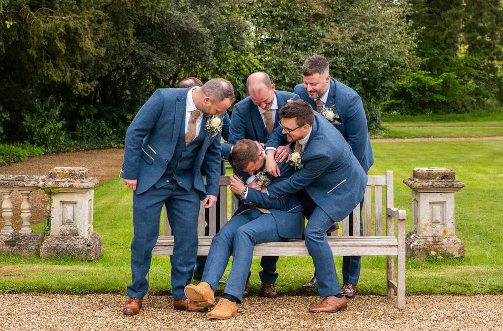 A group of five men in suits, celebrating and laughing together at Orchardleigh House Weddings, one sitting and playfully being checked on by the others outdoors near a bench.
