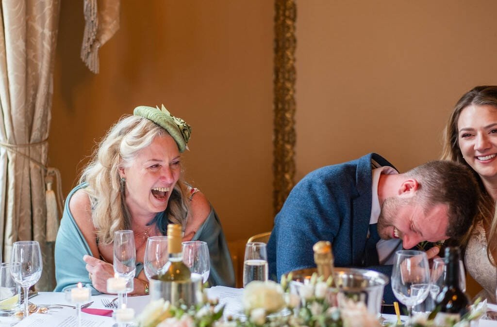 Three people laughing joyously at a dining table during an Orchardleigh House Wedding, with wine glasses and place settings visible.