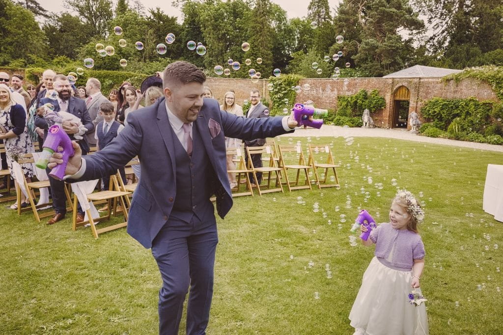 At Elmhay Park, a weddings photographer captures a man and young child in formal attire playfully shooting bubbles into the air, delighting the seated outdoor audience.