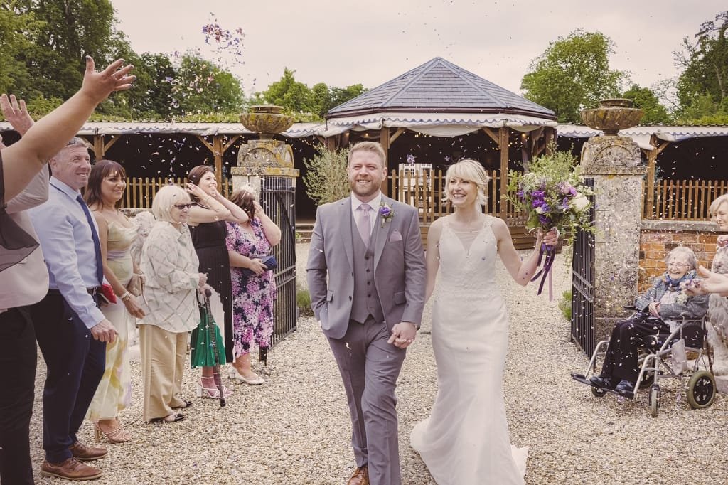 A newly married couple walks down an outdoor aisle at Elmhay Park while guests on both sides throw confetti, capturing the magical moment perfectly for their weddings photographer.