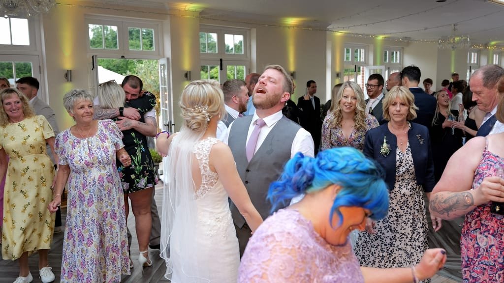 A bride and groom are dancing with guests around them at a wedding reception. Some guests are dancing while others observe. Captured perfectly by the weddings photographer, the venue at Elmhay Park boasts large windows and bright lighting, adding an enchanting touch to the celebration.