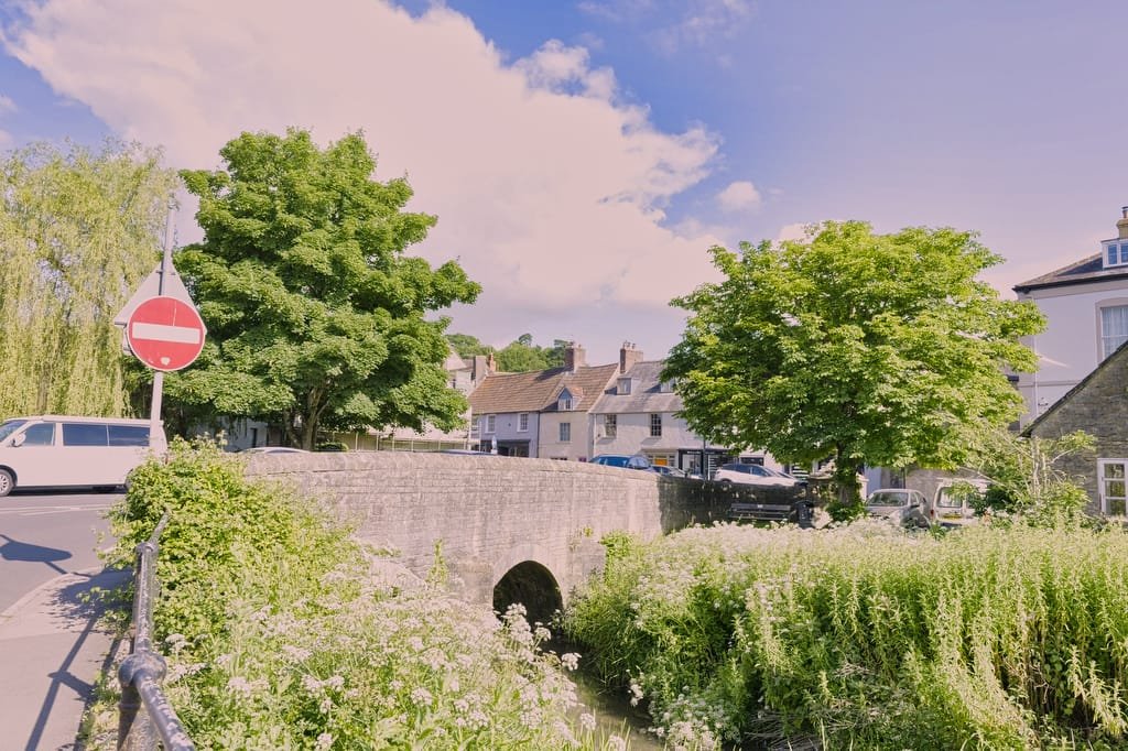 A stone bridge spans a small river in a quaint town, surrounded by greenery and old houses. A "no entry" road sign is visible on the left. The clear, sunny sky provides perfect lighting for a wedding photographer in Bruton.