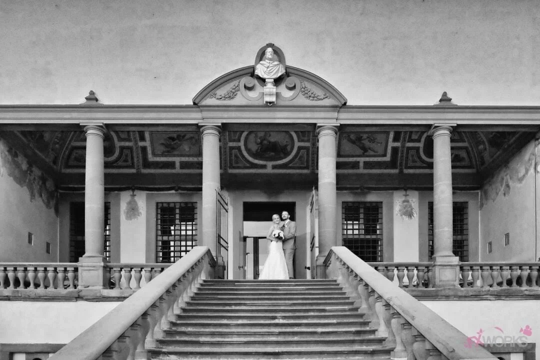 A couple stands at the top of a grand staircase in front of an ornate building, likely on their wedding day. The building, typical of Orchardleigh Weddings, features columns, statues, and decorative elements.