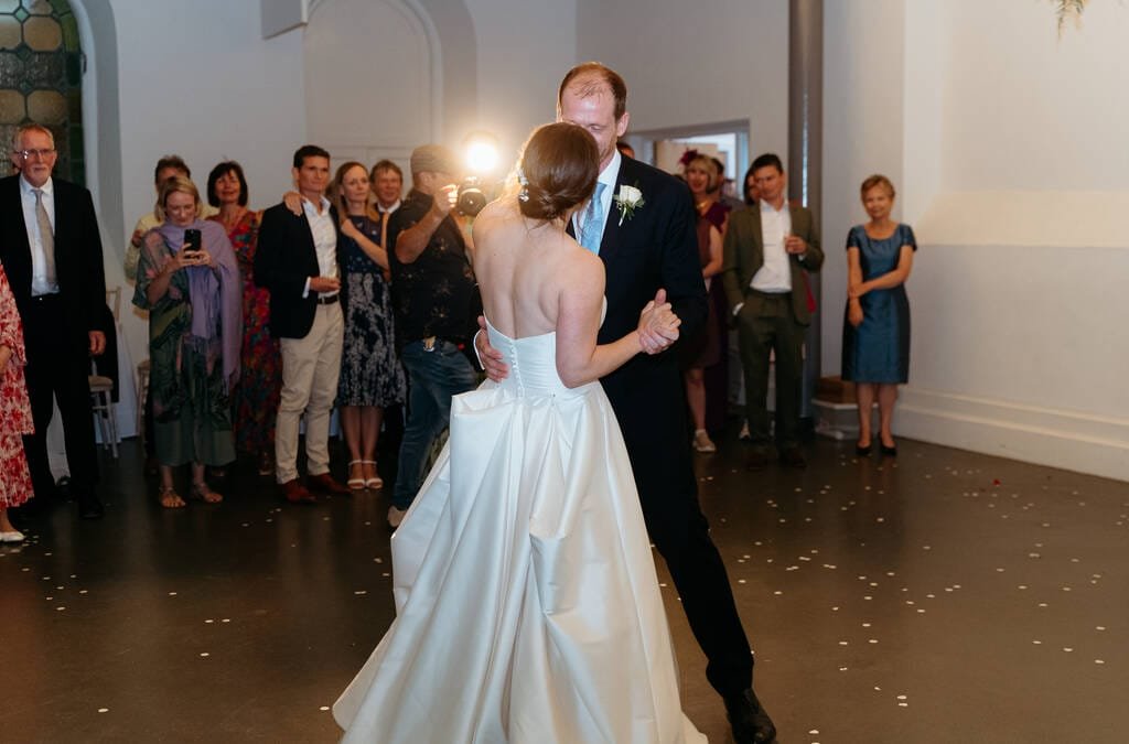 At Rook Lane Weddings in Frome, a bride and groom dance in the center of a room filled with guests, some of whom are taking photographs. The bride wears a white dress, and the groom is in a black suit.