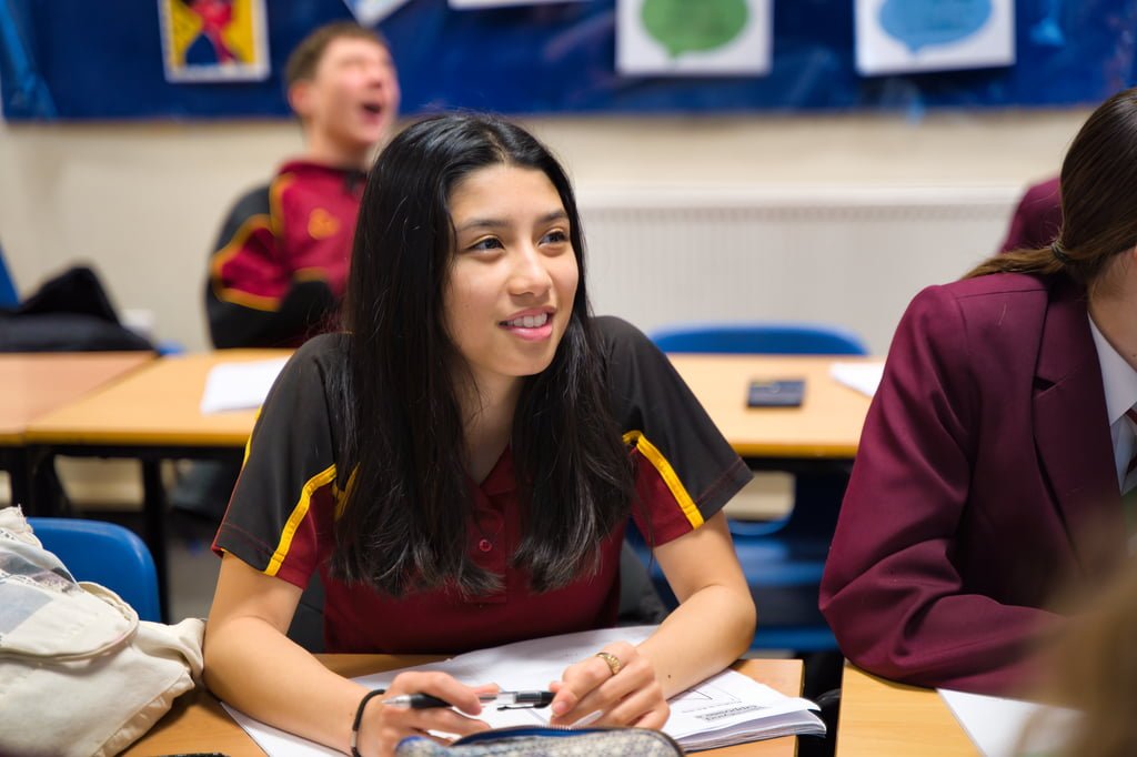 A student in a classroom is engaged in a discussion while seated at a desk with open books and notebooks. Other students are seen in the background, capturing an authentic moment perfect for school marketing photography.