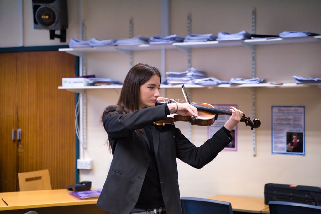 A young woman is standing in a classroom, playing the violin. She is wearing a black blazer, and there are shelves with papers in the background. This image captures a perfect moment of school marketing photography, highlighting both talent and academic environment.