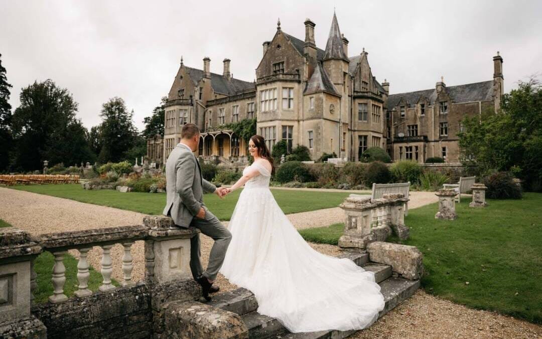 A bride and groom pose on stone steps, framed by the majestic backdrop of Orchardleigh House's historic mansion and manicured gardens.