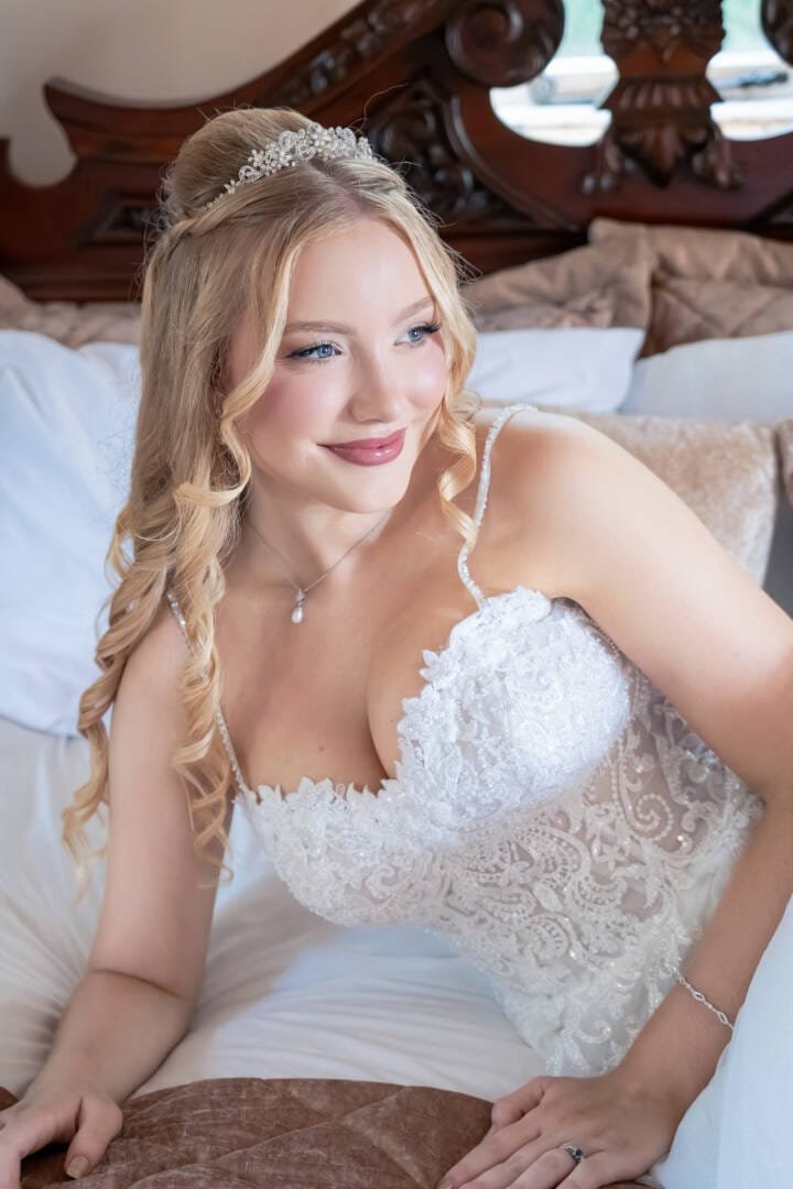 _V5M3357_DxO-Edit - Copy_Original A woman in a white lace wedding dress with long blonde hair and a tiara sits on a bed, smiling and looking to the side, capturing the joy and elegance of Elmhay Park Weddings.