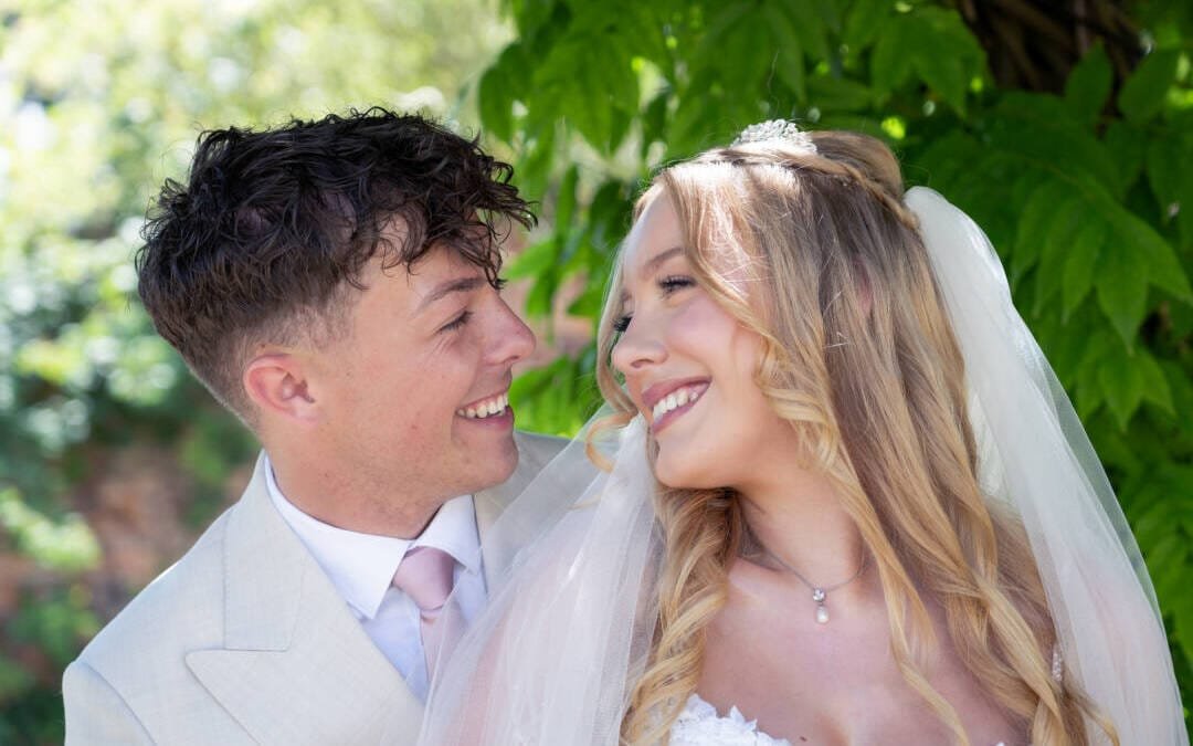 A bride and groom in wedding attire smile at each other outdoors, surrounded by lush greenery—capturing the romance and elegance of Elmhay Park Weddings.