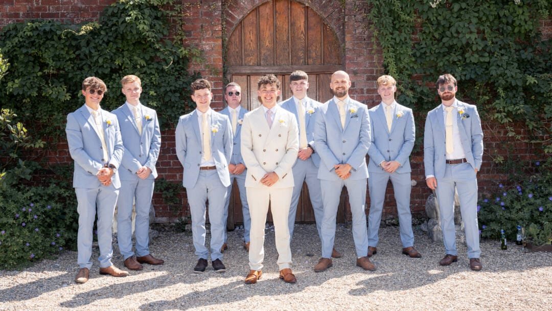_V5M3738_DxO_Original A group of nine men wearing light suits and boutonnieres pose for a photo outdoors in front of a brick wall and arched wooden door, capturing the elegant charm of Elmhay Park Weddings.