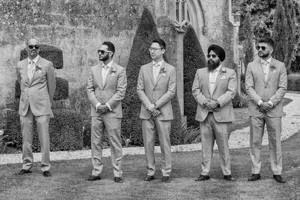 Five men in suits stand in a line outside, in front of trimmed hedges and a stone wall, posing for a photo captured by a Farleigh House Wedding Photographer.