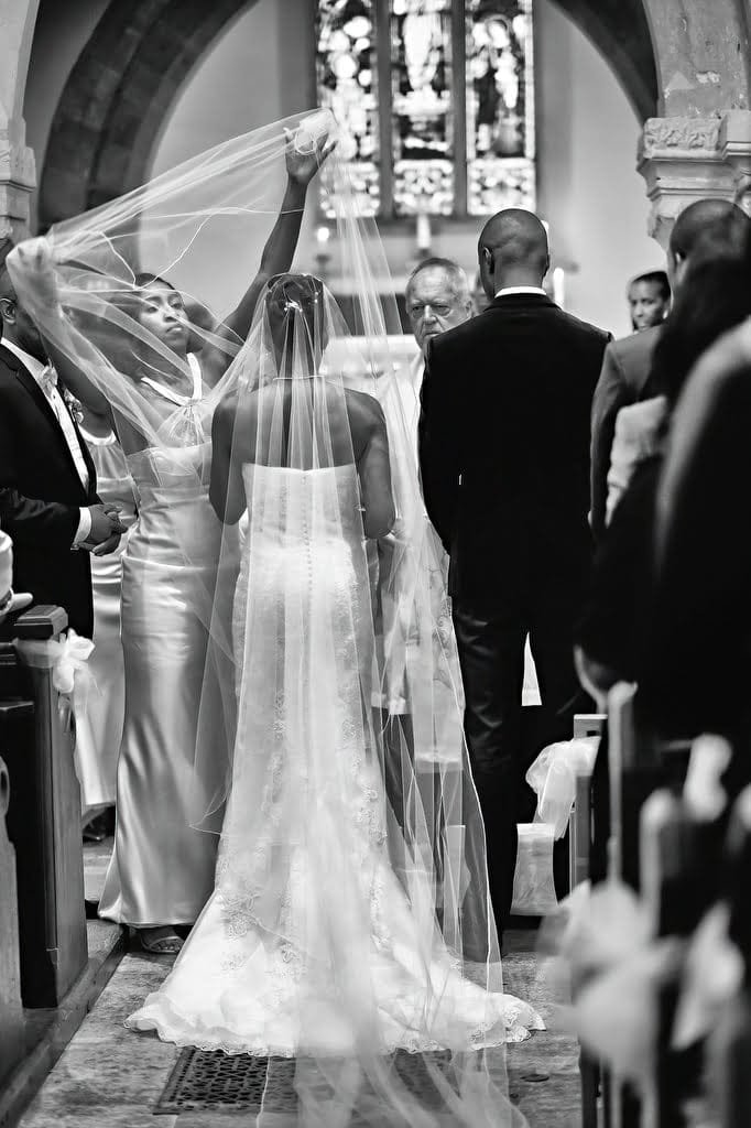 A bride and groom stand at the altar in a church. A woman adjusts the bride's long veil. The scene, reminiscent of an Orchardleigh House Wedding, is in black and white.