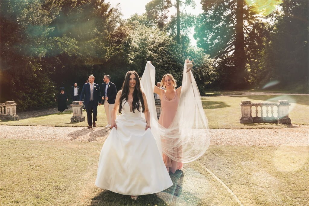 A bride in a white dress walks on a grass path at Orchardleigh House Weddings, with a smiling bridesmaid holding her train. Several people in formal attire walk in the background.