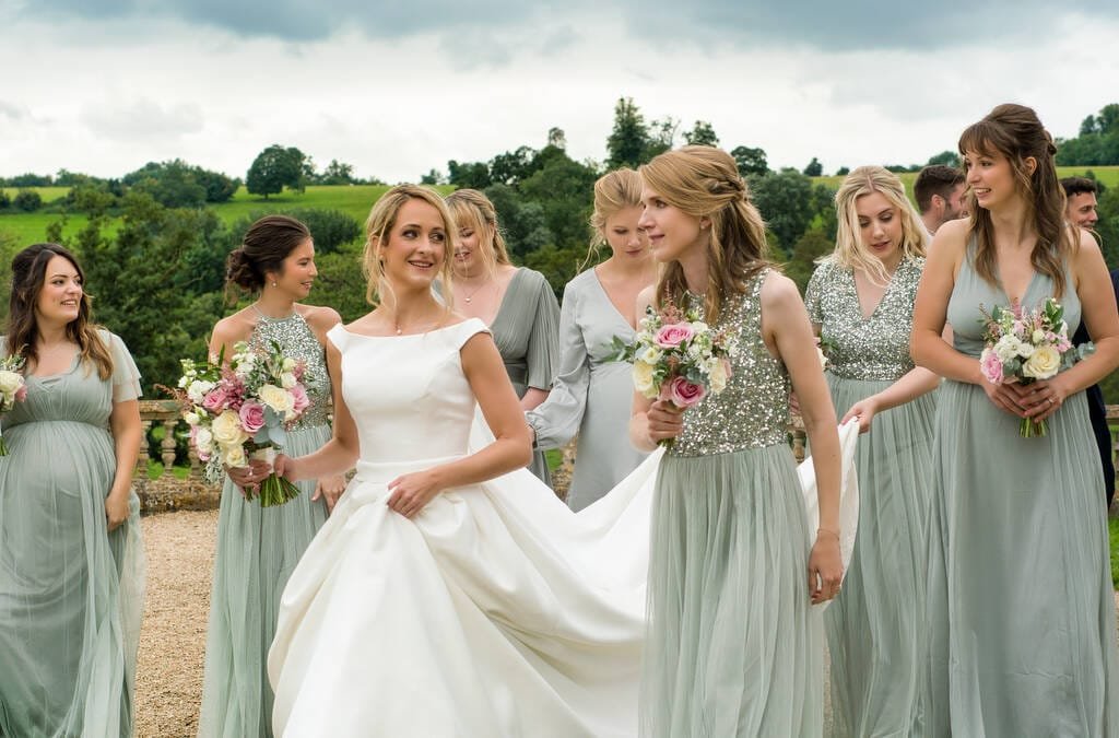 A bride in a white gown is surrounded by bridesmaids wearing light green dresses, holding bouquets. They are outside with a scenic, green landscape in the background, embodying the timeless elegance of Orchardleigh House Weddings.