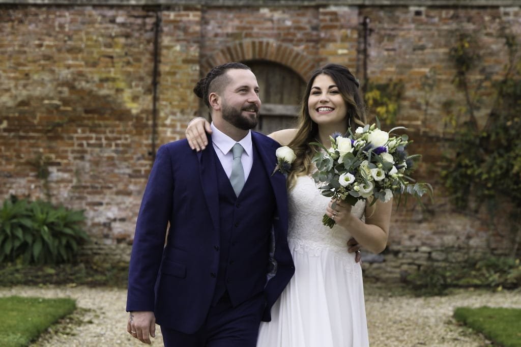 A man in a blue suit and a woman in a white wedding dress holding a bouquet walk arm in arm, smiling, in front of a brick wall with greenery—an enchanting moment reminiscent of Orchardleigh House Weddings.