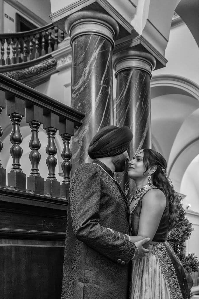 A couple dressed in traditional attire stands closely together, holding hands and gazing at each other, beside ornate columns and a staircase in a grand indoor setting—a beautiful testament to wedding traditions from around the world.