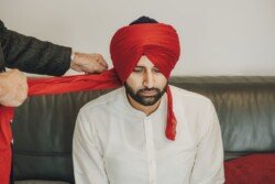 A man dressed in a white shirt is having a red turban tied on his head by another person, creating a picture-perfect moment ideal for capturing with Fuji Cameras for weddings.