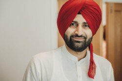 A man wearing a red turban and white traditional attire smiles while looking at the camera, captured beautifully with Fuji Cameras for weddings.
