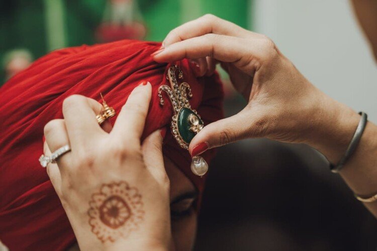 A close-up of hands adjusting a decorative headpiece on a red turban conveys the rich wedding traditions from around the world. The person has a ring on one finger and a henna design on the back of their hand, symbolizing cultural beauty and commitment.