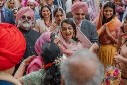 A group of people in traditional attire, including turbans and colorful dresses, gather outdoors near the historic Guildhall. They look happy and engaged in conversation. A woman in the center smiles broadly, creating an atmosphere as warm as a summer day in Bath.