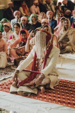 A seated person in traditional wedding attire, including a turban and face veil, holds a staff, surrounded by attendees dressed in colorful clothing inside a ceremony hall—a scene beautifully captured by Fuji Cameras for weddings.