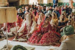 A couple wearing traditional attire sits on the floor during a Sikh wedding ceremony, surrounded by attendees. The bride, in a red dress, and the groom, in a white outfit with a turban, create perfect moments for capturing with Fuji Cameras for weddings.