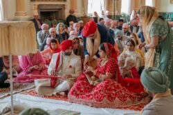 A couple dressed in traditional attire sit together during a wedding ceremony at the Guildhall, surrounded by guests wearing colorful garments, in a large decorated room.