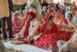 A bride in a red and gold outfit and a groom in white and red attire sit on the floor among family and friends during a traditional wedding ceremony at the elegant Guildhall Bath.