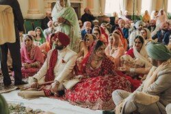 A couple in traditional attire takes part in a ceremony. The bride wears a red dress, and the groom has a red turban. Numerous people dressed in colorful clothes are seated around them, capturing the beautiful moments with their Fuji Cameras for weddings.