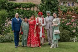 A group of six people stands in front of a garden, reminiscent of the picturesque scenes at Guildhall Bath. Four of them wear traditional South Asian attire, while the other two are dressed in Western-style formal wear. They pose together smiling.