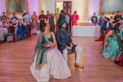 A couple in traditional Indian attire sits smiling at an indoor event, surrounded by guests in the elegant Guildhall. The venue, adorned with paintings on the walls and a polished wooden floor, exudes a sense of timeless charm.
