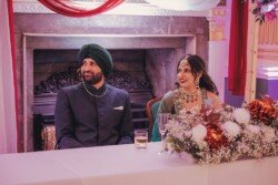 A couple in traditional attire sits at a table adorned with floral arrangements, in front of an ornate fireplace background at a formal event, perfectly captured by Fuji Cameras for weddings.