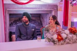 A man in a turban and a woman in traditional attire sit at a decorated table with flowers, in front of a fireplace at what appears to be a formal event or ceremony, perhaps within the historic ambiance of the Guildhall Bath.