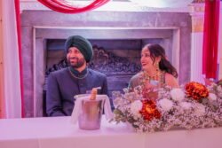 A man and woman are seated at a decorated table near a fireplace in the charming Guildhall Bath, both smiling, dressed in traditional attire. The table boasts floral arrangements and a bucket with a bottle.