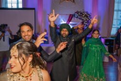 People dressed in traditional clothing are dancing energetically at a festive event held at the historic Guildhall Bath, with beautiful floral decorations in the background.