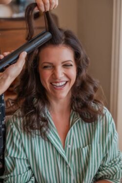 A woman with curly hair smiles while another person styles her hair with a flat iron at Orchardleigh House. She is wearing a green and white striped shirt.
