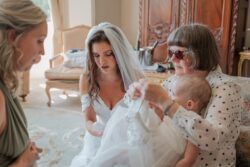 A bride in a white dress and veil stands beside an elderly woman in sunglasses, who is dressing a baby in a white outfit. Another woman with blonde hair sits nearby. Set against the backdrop of antique furniture, this intimate moment unfolds within the elegant surroundings of Orchardleigh House.