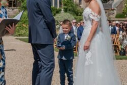A young boy in a blue suit holds up a ring box during an outdoor wedding ceremony at Orchardleigh House. The bride and groom stand facing each other, surrounded by guests seated on wooden chairs.
