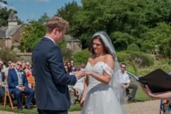 A bride and groom exchange rings during an outdoor wedding ceremony at Orchardleigh House, with guests seated in the background. The bride is in a white dress and veil, while the groom wears a dark suit.