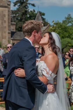 A bride and groom kiss during an outdoor wedding ceremony at Orchardleigh House while guests look on. The bride is in a white dress and veil, and the groom is in a dark suit. Trees and the elegant building of Orchardleigh House are visible in the background.