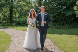 A bride in a white dress and groom in a suit walk together on a gravel path surrounded by greenery at Orchardleigh House, smiling.