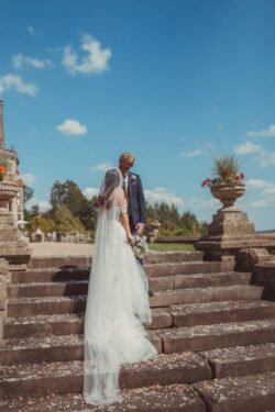 A bride and groom in formal attire stand close together on the stone steps of Orchardleigh House, surrounded by scattered flower petals, with a clear blue sky in the background.