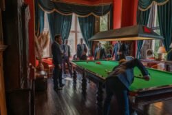 A group of men in suits stand around a pool table in a room at Orchardleigh House, with red walls, large windows, and dark green curtains. One man is bent over the table, preparing to take a shot.