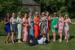 A group of 12 people, mostly women in colorful dresses, poses for a photo in the grassy outdoor setting of Orchardleigh House. One man in a suit lies playfully on the ground in front.