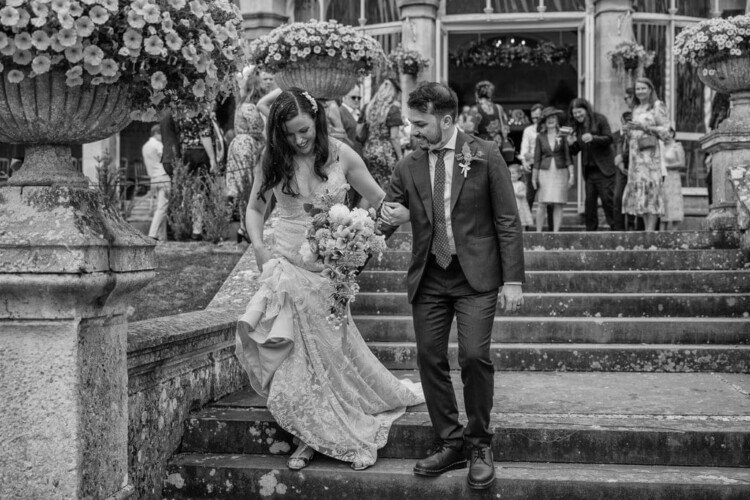 A bride and groom, holding hands, walk down a set of stone steps at Grittleton House. The bride holds a bouquet of flowers, as guests stand in the background near the grand building, captured perfectly by the photographer.