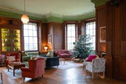 A spacious living room with wood paneling, ideal for Hartham Park Weddings. It features a decorated Christmas tree, several colorful armchairs, a sofa, and large windows that fill the space with light.