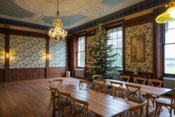 The decorated room at Hartham Park Weddings features wooden tables and chairs, a sparkling Christmas tree, elegant chandeliers, floral wallpaper, and large windows that flood the space with natural light.