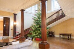 A large Christmas tree, perfect for Hartham Park Weddings, is adorned with lights and ornaments and stands gracefully in a spacious room with a wooden staircase and columns. Wrapped gifts are elegantly placed beneath the tree.